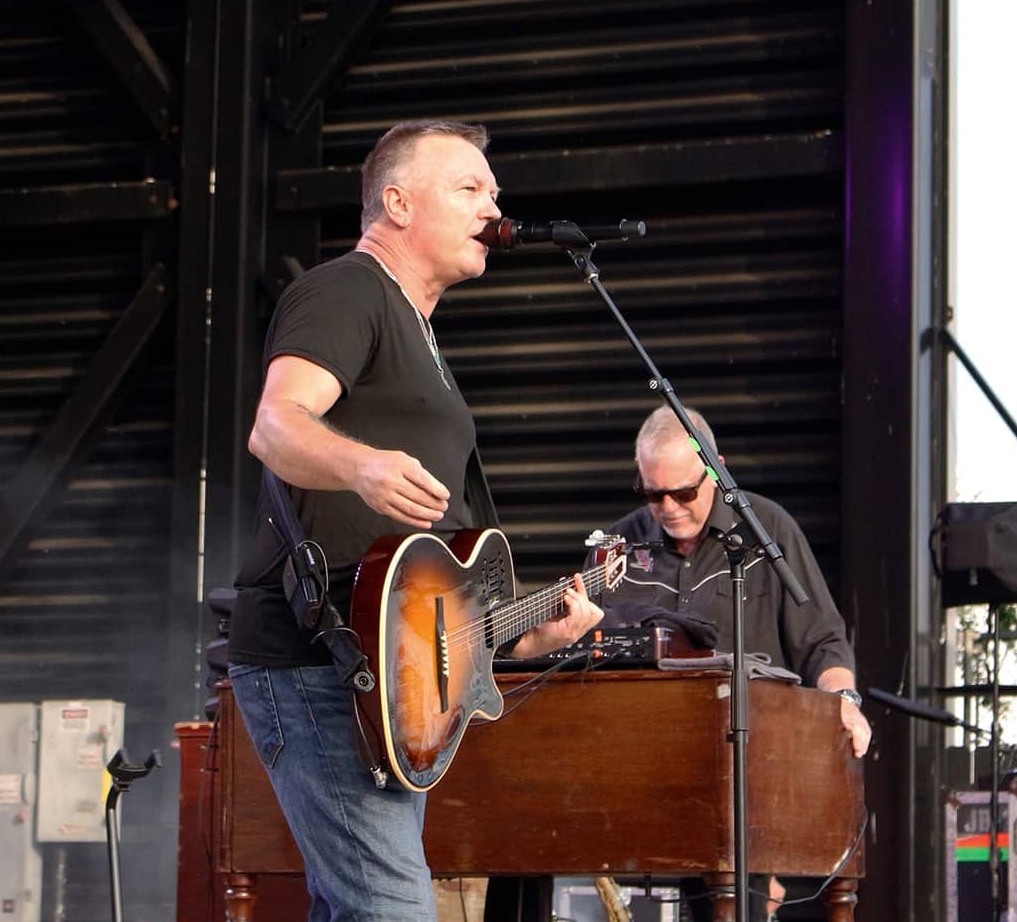 Edwin McCain sings and plays guitar on stage while another plays keyboard in the background. Both are wearing black shirts. The stage has equipment and lighting behind them.