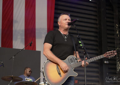 A man sings into a microphone while playing an acoustic guitar on stage. Behind him, a drummer plays near a large American flag. Stage lights and equipment are visible in the background.