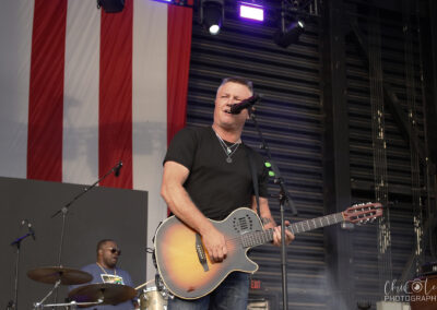 A musician plays an acoustic guitar and sings into a microphone on an outdoor stage, with a drummer performing behind him and a large American flag hanging in the background.