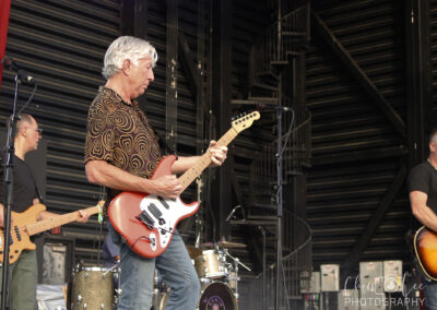 A band performs on stage with three guitarists playing electric guitars and a drum set visible in the background. The musicians are focused on their instruments. The stage is set with large black panels.