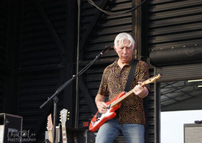 An older man with gray hair plays an electric guitar on stage. He wears a patterned shirt and jeans, with additional guitars visible nearby. The setting appears to be an outdoor or open-air venue.