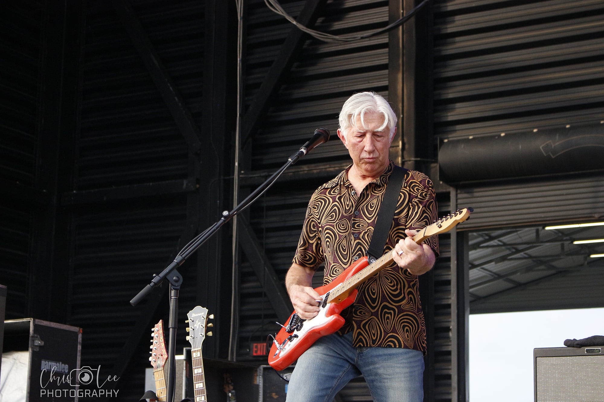 An older man with gray hair plays an electric guitar on stage. He wears a patterned shirt and jeans, with additional guitars visible nearby. The setting appears to be an outdoor or open-air venue.