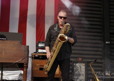 Male saxophonist wearing sunglasses and a black shirt performs on stage with a red-and-white striped backdrop.