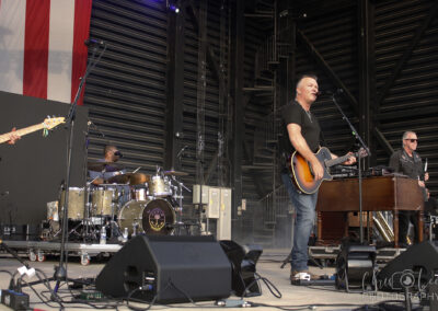 A band performs on an outdoor stage; the lead singer plays an acoustic guitar and sings into a microphone, with a drummer and keyboard player in the background under a large American flag.