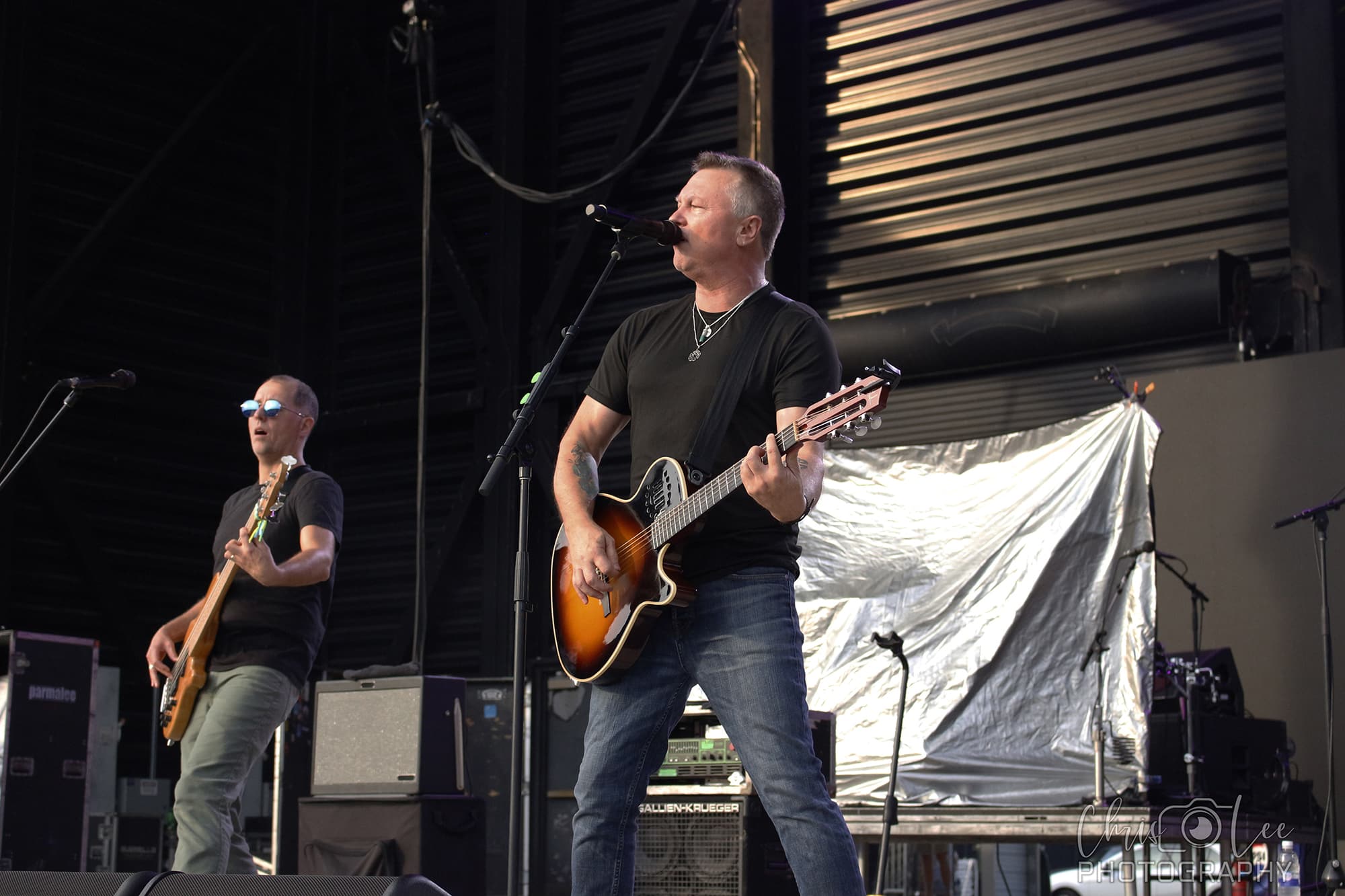Two musicians perform on stage; one sings and plays an acoustic guitar while the other plays bass. Both wear dark shirts and sunglasses. Stage equipment and a metallic backdrop are visible behind them.