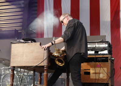 A musician in sunglasses plays a keyboard on stage, with a saxophone hanging at his side. Smoke rises behind him, and a large American flag hangs in the background. Music equipment is visible around him.