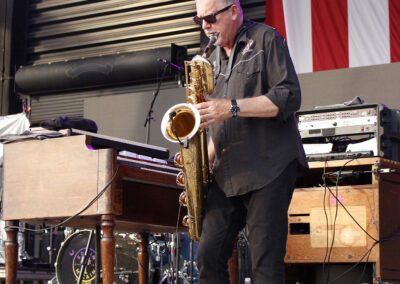 Male saxophonist wearing sunglasses performing on stage with purple lights and a large American flag backdrop behind him.