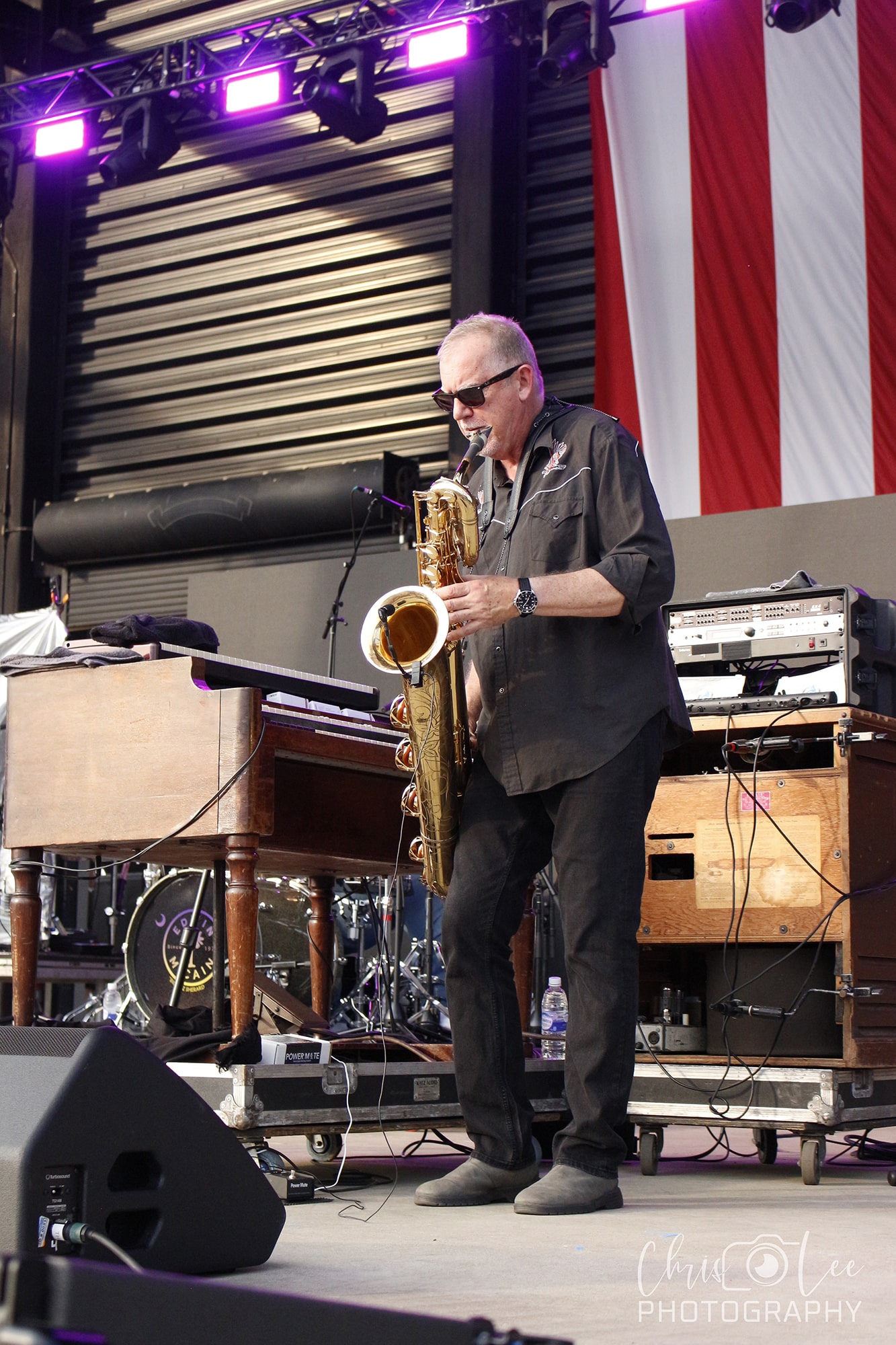 Male saxophonist wearing sunglasses performing on stage with purple lights and a large American flag backdrop behind him.