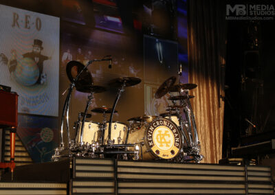 A Kevin Cronin Band drum set on a stage platform, featuring a bass drum logo against a backdrop of REO Speedwagon album art at the PNC Pavilion.