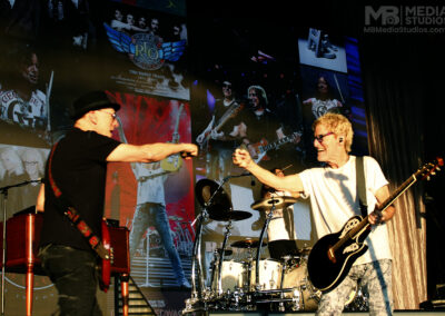 Two Kevin Cronin Band members fist-bump while performing on stage at the PNC Pavillion in front of a large screen displaying archival photos of the band.