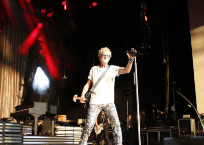 Kevin Cronin with blonde hair and glasses singing into a microphone on stage while holding an electric guitar at the PNC Pavilion.