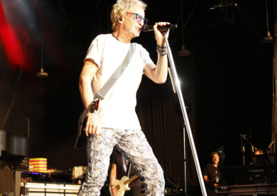 Kevin Cronin sings into a microphone stand on the Kevin Cronin Band stage, wearing a white t-shirt and patterned pants.