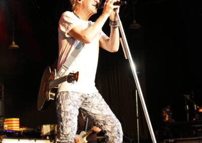 Kevin Cronin sings into a microphone on stage at the PNC Pavilion while holding an acoustic guitar.