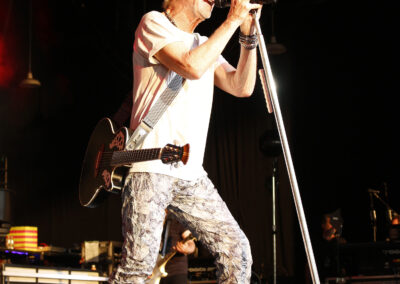 Kevin Cronin sings into a microphone while holding an acoustic guitar on stage during the Brotherhood of Rock Tour.