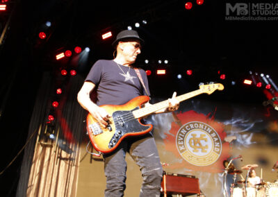 Matt Bissonette wearing a fedora and sunglasses plays a sunburst bass guitar on stage in front of a Kevin Cronin Band backdrop during the Brotherhood of Rock Tour.