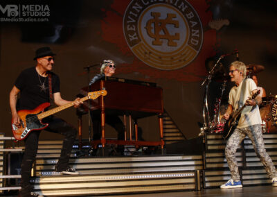 The Kevin Cronin Band performing live on stage at the PNC Pavilion with a bassist, a keyboardist, and a guitarist.