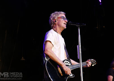 Kevin Cronin performs with an acoustic-electric guitar at a microphone on a dark stage while wearing colorful glasses.