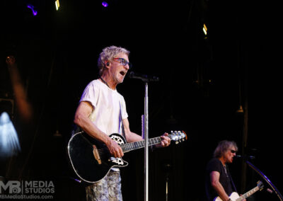 Kevin Cronin sings and plays a black acoustic-electric guitar on stage during the Brotherhood of Rock Tour, with a bassist visible in the background.