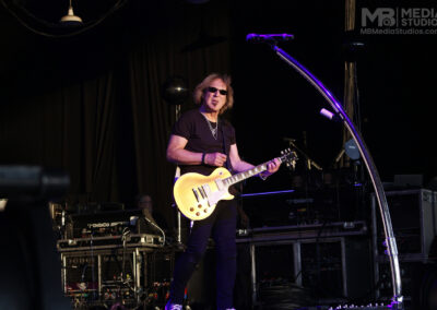 A musician with long brown hair and sunglasses plays a yellow electric guitar on stage with the Kevin Cronin Band.