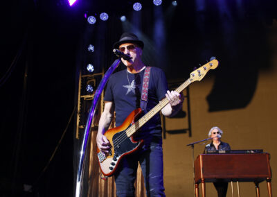 A musician in a fedora and a star-print t-shirt plays a sunburst bass guitar and sings into a microphone on the Brotherhood of Rock Tour stage, with a keyboardist performing in the background.