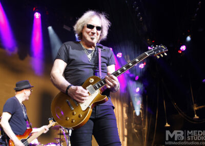 A guitarist wearing sunglasses and a black t-shirt plays a gold-top electric guitar on stage at the PNC Pavilion, with a bass player visible in the background.