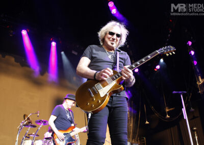 Guitarist with wavy hair and sunglasses, plays a gold-top electric guitar on stage at PNC Pavilion while a bassist performs in the background.