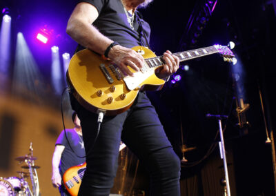 A guitarist with curly hair and sunglasses plays a yellow electric guitar on stage during the Brotherhood of Rock Tour, with a bassist visible in the background.