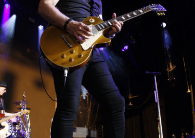 A long-haired guitarist wearing sunglasses plays a gold-top electric guitar on stage under concert lights during the Brotherhood of Rock Tour.