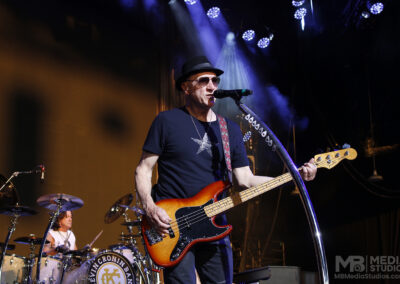 Matt Bissonette sings and plays a sunburst electric bass guitar on stage at the PNC Pavilion, with a drummer visible in the background.