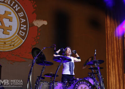 A drummer sings into a microphone while performing on stage during the Brotherhood of Rock Tour in front of a large Kevin Cronin Band logo backdrop.