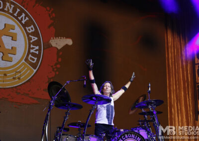 A drummer with arms raised behind his kit at the PNC Pavillion in front of a large screen featuring the Kevin Cronin Band logo.