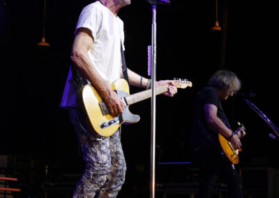 Kevin Cronin sings into a microphone while playing an electric guitar at the PNC Pavilion, with a guitarist performing in the Brotherhood of Rock background.