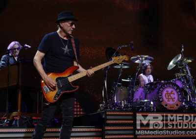 A bassist wearing a hat and black t-shirt performs on the Brotherhood of Rock Tour alongside a keyboardist and a drummer with a branded drum kit at the PNC Pavilion.