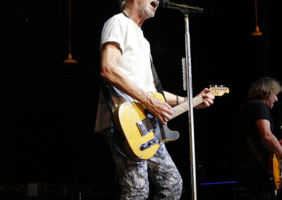 Kevin Cronin singing into a microphone while playing an electric guitar on the Kevin Cronin Band stage.