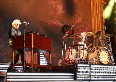 A keyboardist playing an organ and a drummer performing with the Kevin Cronin Band on stage in front of a large, glowing red backdrop.